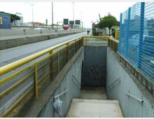 Sottopassaggio pedonale con scala in cemento che scende sotto una strada a scorrimento veloce, affiancata da una ringhiera gialla e una recinzione metallica blu. In fondo alla rampa si vede un muro con graffiti, mentre lungo la carreggiata ci sono cartelli stradali e infrastrutture urbane.
