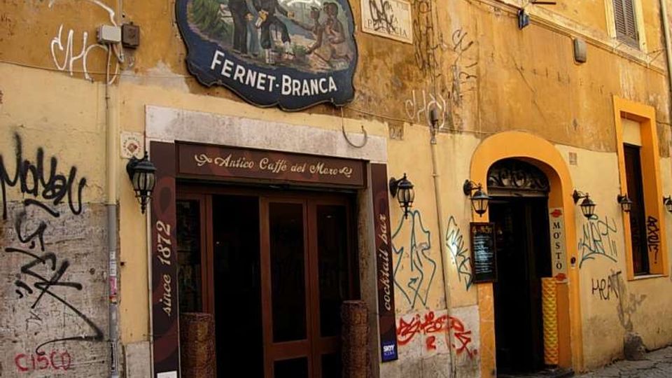 Facciata di un edificio storico con intonaco giallo e numerosi graffiti, con insegna vintage “Antico Caffè del Moro – Fernet-Branca” sopra l’ingresso. Due porte ad arco con lanterne a parete si affacciano su una strada lastricata in pietra.