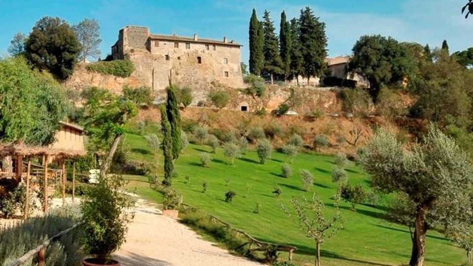 Veduta di una collina con un grande edificio in pietra tipo castello o villa fortificata, circondato da cipressi e alberi. In primo piano un viale sterrato e un giardino con prato verde e ulivi.