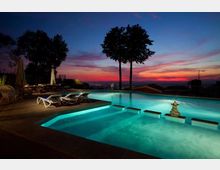Piscina illuminata all’aperto con gradini e sedute sommerse, circondata da sdraio e ombrelloni su una terrazza. Sullo sfondo si vedono alberi in controluce e una vista panoramica verso l’orizzonte al tramonto.