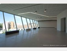 Ampia sala interna moderna e vuota, con pavimento chiaro e soffitto a pannelli con illuminazione lineare e faretti. Sul lato sinistro una lunga parete di grandi vetrate inclinate offre vista su edifici urbani e fa entrare molta luce naturale.