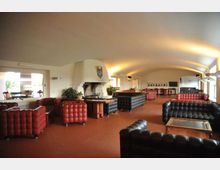Ampio salone interno con soffitto a volta illuminato, pavimento in piastrelle e grande camino in muratura su una parete. Sono presenti divani Chesterfield in pelle rossa e nera disposti in gruppi con tavolini, con finestre laterali e area bar in fondo alla sala.