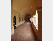 Corridoio di un chiostro in pietra con soffitto a volta e pavimento in mattoni disposti a spina, illuminato da una fila di arcate e colonnine sul lato destro. Sul lato sinistro si vedono porte e nicchie ad arco nella muratura, con lampade a sospensione lungo il percorso.