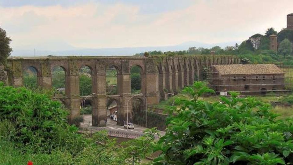 Grande acquedotto in pietra con una lunga serie di arcate che attraversa una valle verde, con una strada che passa sotto gli archi. Sulla destra si vedono un edificio rurale con tetto in tegole e una torre cilindrica su una collina alberata sullo sfondo.