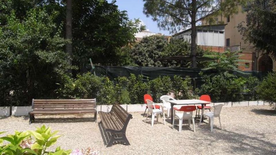 Cortile-giardino con area in ghiaia, due panchine in legno e un tavolo con sedie in plastica bianche e rosse. Sullo sfondo ci sono alberi e arbusti fitti, con un edificio chiaro parzialmente visibile dietro la vegetazione e una rete di recinzione.