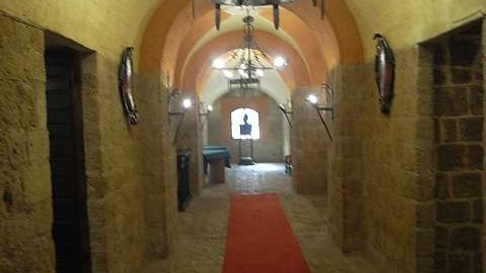 Corridoio sotterraneo in pietra con soffitto a volta e pavimento in mattoni, illuminato da grandi lampadari in ferro battuto. Un tappeto rosso corre lungo il centro fino a un arco in fondo, con nicchie e aperture laterali nelle pareti.