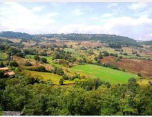 Panorama di campagna collinare con campi coltivati a mosaico e aree boschive, attraversate da strade e piccoli casali sparsi. In lontananza si vedono dorsali collinari sotto un cielo parzialmente nuvoloso.