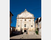 Facciata di una chiesa storica in stile barocco, con frontone curvilineo decorato, finestre ovali laterali e ingresso centrale sopra una scalinata in pietra con cancellata in ferro. La chiesa è affiancata da edifici residenziali e attraversata da cavi sospesi nel cielo azzurro.
