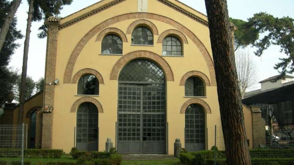 Facciata di un edificio in stile industriale con intonaco giallo e archi in mattoni, con grande portale centrale vetrato e finestre ad arco simmetriche. Davanti si vede un prato con siepi basse e alberi alti che incorniciano l’ingresso.