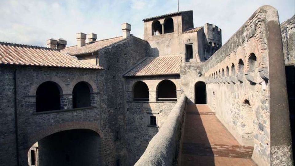Cortile interno di un edificio storico in pietra con tetti in tegole, arcate su più livelli e un camminamento sopraelevato con parapetto e pavimento in cotto. Sulla destra si vede un lungo muro con una fila di piccole aperture e un accesso ad arco sul fondo.