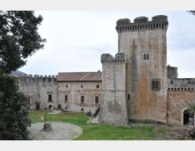 Cortile di un castello in pietra con alte torri merlate e mura fortificate, affiancate da un edificio con tetto in tegole e finestre rettangolari. In primo piano si vede un prato con un’area circolare in ghiaia e un pozzo, con un arco d’ingresso sulla destra.