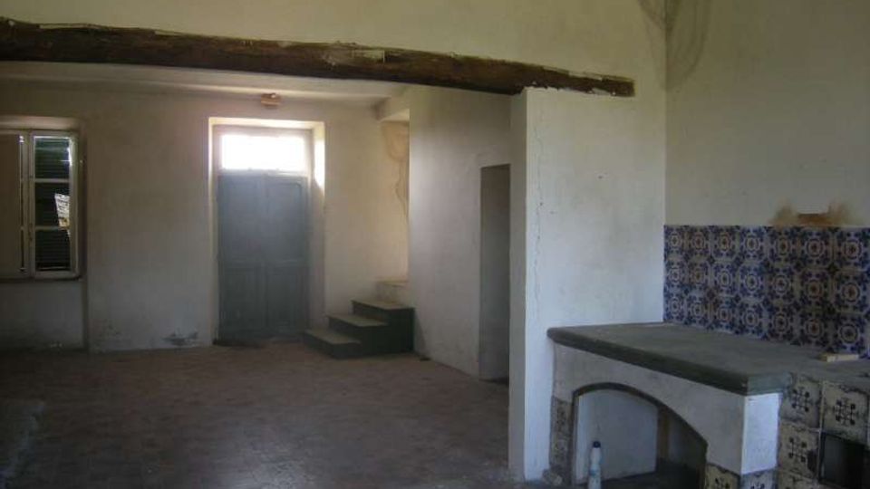 Interno di una vecchia cucina rurale con pavimento in mattoni, travi a vista e pareti intonacate. A destra c’è un grande banco in muratura rivestito di piastrelle decorative, mentre sul fondo si vedono una porta, una finestra e un piccolo gradino che conduce a un vano.