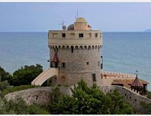 Torre costiera in pietra di forma cilindrica con merlature e terrazza superiore, collegata da una rampa a un complesso con mura e piccoli tetti in tegole. Sullo sfondo si vede il mare, con vegetazione bassa e alberi in primo piano.