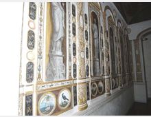 Parete interna di un edificio storico decorata con affreschi e stucchi dorati, con nicchie ad arco contenenti figure monocrome e pannelli ovali con paesaggi e uccelli. Un corridoio stretto corre lungo la parete, con luce intensa proveniente da sinistra.