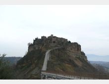 Borgo e complesso di edifici in pietra su una rupe rocciosa, collegati da una lunga strada sopraelevata che sale fino all’ingresso. Sullo sfondo si vedono colline e una valle sotto un cielo coperto.