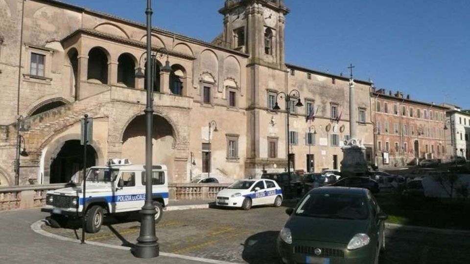 Piazza urbana con pavimentazione in pietra e auto parcheggiate, affacciata su un grande edificio storico in muratura con porticato ad archi, scala esterna e torre campanaria con orologio. Lampioni in ferro e facciate di palazzi allineati lungo la strada sotto un cielo sereno.