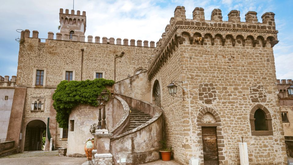 Cortile di un castello in pietra con merlature e torre, affacciato su una facciata con finestre ad arco e portale. Una scalinata esterna curva sale lungo il muro, con lampioni e una zona coperta di edera vicino all’ingresso.