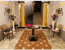 Ingresso interno in stile classico con pavimento in marmo e riquadri in cotto, tavolino rotondo centrale e due colonne ocra che incorniciano una scala in fondo. Lungo le pareti grigie con cornici gialle ci sono sedie, specchi e alti vasi con composizioni floreali, sotto un soffitto ad arco decorato e lampadario.