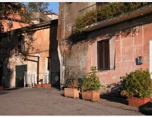 Cortile esterno con pavimentazione in cemento affacciato su edifici intonacati in rosa e grigio, con finestre con persiane, porta verde e muri segnati dal tempo. In primo piano ci sono grandi fioriere con arbusti e, vicino all’ingresso, una pila di sedie bianche impilate.