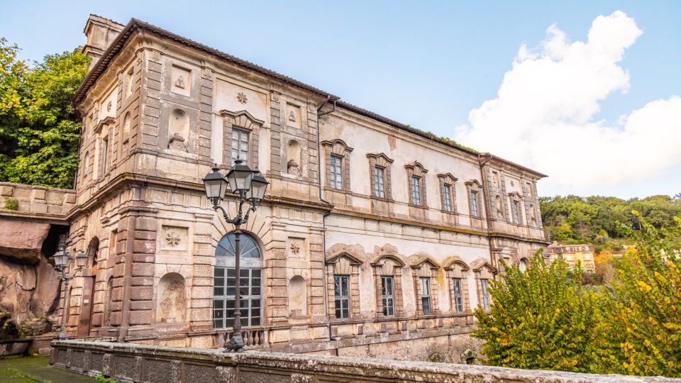 Edificio storico in pietra e intonaco, a due piani, con facciata decorata da cornici e nicchie e una grande porta ad arco con vetrate. In primo piano si vede un parapetto in pietra e un lampione a più bracci, con alberi e colline boscose sullo sfondo.