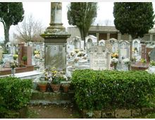 Cimitero con numerose tombe e lapidi in pietra disposte in file, alcune decorate con fiori e vasi. In primo piano si vede un monumento funerario con base in pietra accanto a siepi basse, con alberi alti sullo sfondo.
