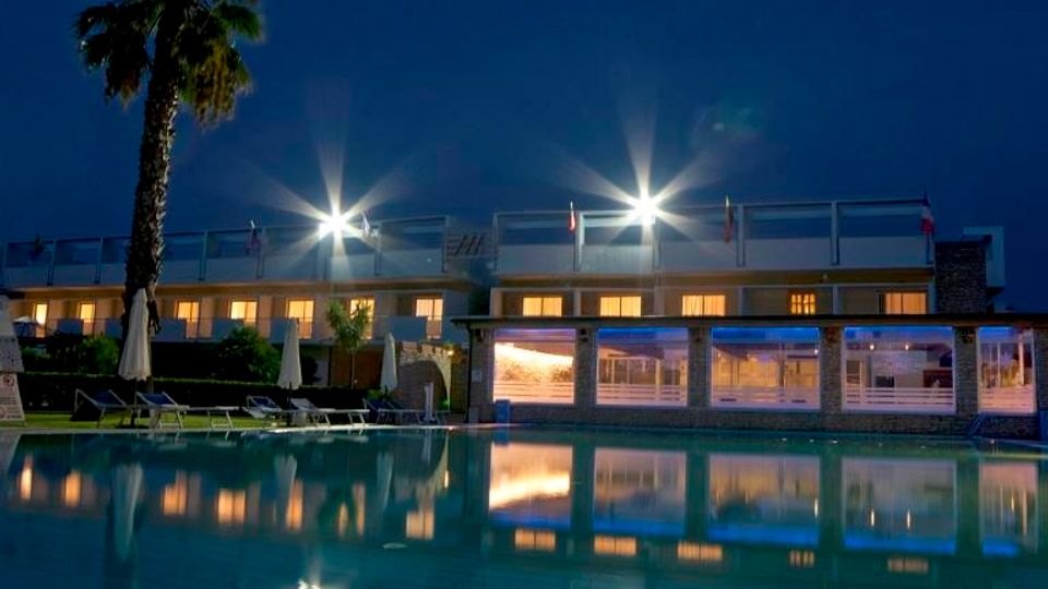 Piscina esterna di un hotel al crepuscolo, con acqua calma che riflette le luci dell’edificio moderno a due piani. A bordo piscina si vedono lettini e ombrelloni chiusi, una palma e grandi vetrate illuminate sul lato della struttura.