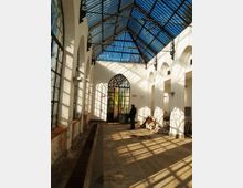 Interno di una galleria coperta con tetto in vetro e struttura metallica, pareti bianche con arcate e grandi finestre laterali. In fondo si vede un portale ad arco vetrato, con luce solare che proietta ombre geometriche sul pavimento.