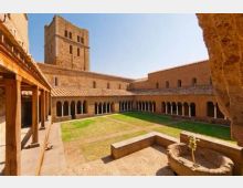 Chiostro di un complesso monastico in pietra con portici ad arcate su tre lati, cortile centrale erboso e un antico pozzo in primo piano. Sullo sfondo si vede una torre campanaria quadrata e edifici con tetti in tegole sotto un cielo sereno.