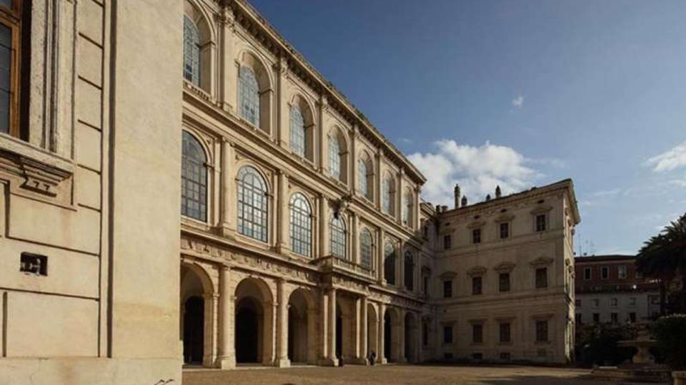 Cortile interno di un palazzo storico in pietra, con loggiato ad archi al piano terra e grandi finestre ad arco sui livelli superiori. Lo spazio è ampio e pavimentato, circondato da facciate classiche sotto un cielo sereno.