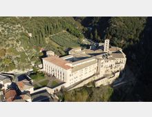 Veduta aerea di un grande complesso monastico in pietra con tetti in coppi rossi, chiostro interno e campanile, affacciato su una scarpata. Intorno si vedono una valle boscosa, campi coltivati e piccole strade di accesso.