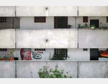 Facciata di un edificio residenziale in cemento con lunghi balconi sovrapposti, ringhiere piene e piccole finestre a griglia. Sui balconi si vedono vasi con piante e un murale/graffito colorato su una parete.