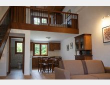 Interno di un appartamento a pianta aperta con soppalco in legno e ringhiera in metallo, scala laterale e soffitto con travi a vista. Al piano inferiore ci sono cucina e zona pranzo con mobili in legno e grandi finestre sul verde, un bagno visibile dalla porta a sinistra e un soggiorno con divano e credenza.