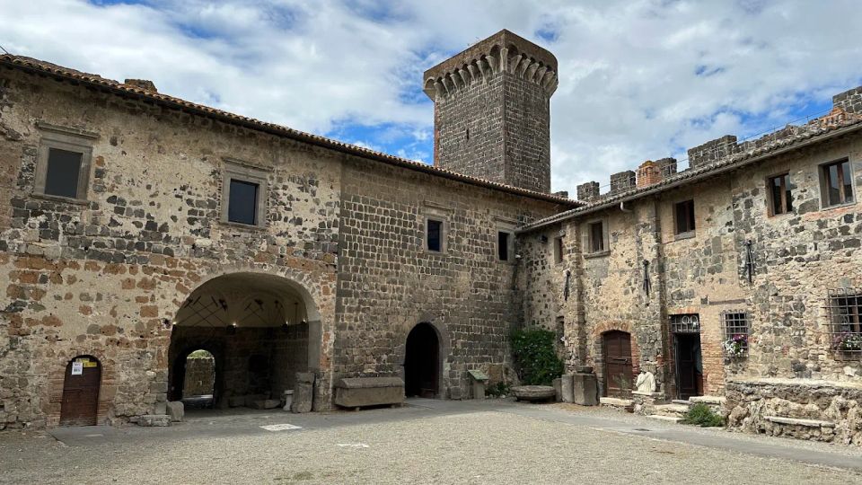 Cortile interno di un castello in pietra, circondato da edifici con muri in tufo e mattoni, finestre piccole e portali ad arco. Sullo sfondo si vede una torre quadrata merlata e un camminamento con merli sotto un cielo parzialmente nuvoloso.