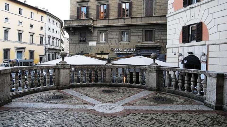 Terrazza urbana pavimentata a motivi geometrici in pietra e mattoni, delimitata da una balaustra in pietra con colonnine e pilastri. Sullo sfondo si vedono palazzi storici con persiane e finestre e alcune coperture bianche di dehors su una strada cittadina.