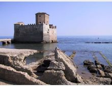 Castello in pietra affacciato sul mare, con torre centrale e mura merlate, costruito su un piccolo isolotto vicino alla riva. In primo piano si vedono scogli e rovine in pietra lungo la costa e acque calme sotto un cielo sereno.