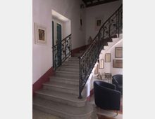 Interno di un edificio con scala in pietra che sale su due rampe, dotata di ringhiera in ferro battuto decorata. Pareti bianche con fascia rossa, quadri e cornici appesi, e un piccolo angolo salotto con poltrone e tavolino accanto alla scala.