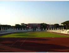 Vista frontale di uno stadio con pista di atletica rossa che circonda un campo in erba verde. Sullo sfondo si vedono gradinate in pietra con statue allineate e un edificio principale in mattoni rossi, circondato da alberi.