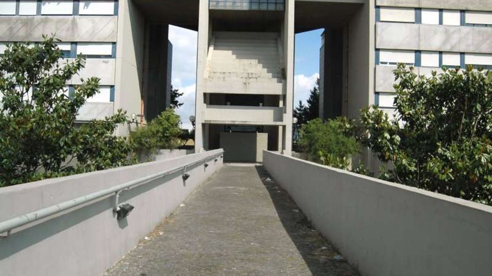 Passerella pedonale in cemento con corrimano che conduce all’ingresso di un grande edificio moderno in calcestruzzo, con ampie aperture centrali e finestre a griglia. Vegetazione e alberi fiancheggiano il percorso ai lati.