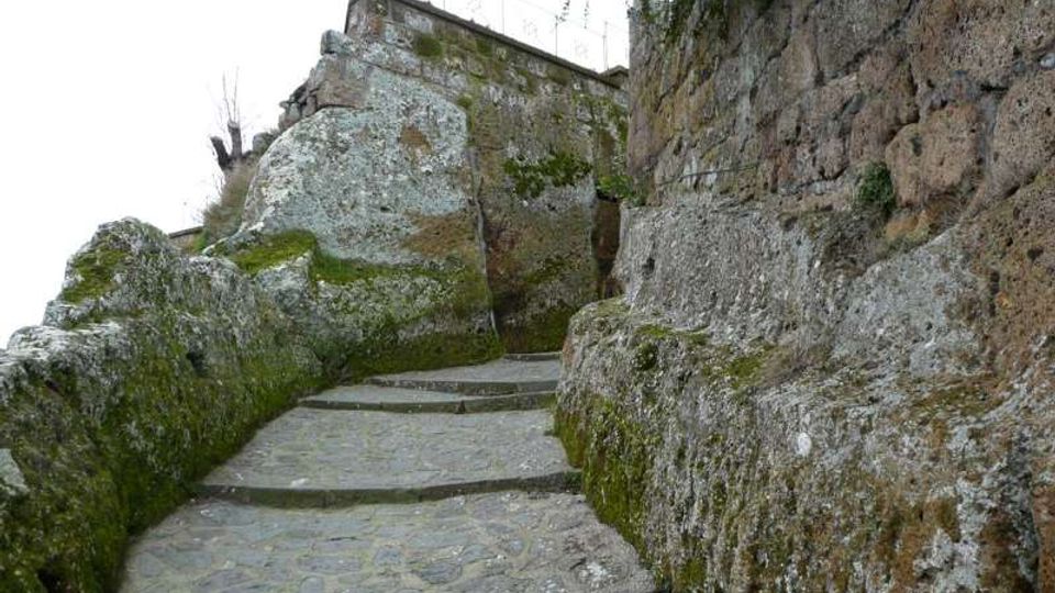 Sentiero in pietra a gradoni che sale tra grandi rocce e una parete rocciosa, con muschio e licheni sulle superfici. In alto si vede un muretto in muratura con ringhiera metallica lungo il bordo.