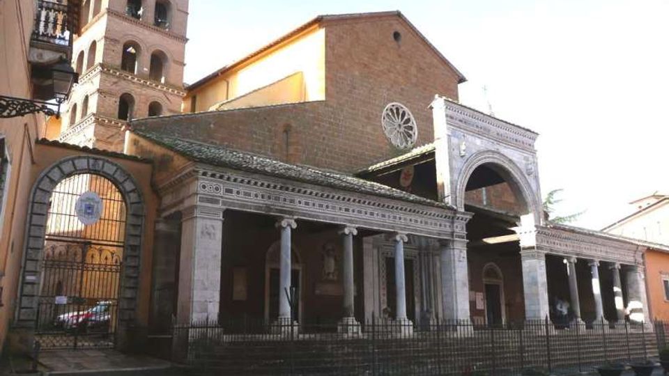 Facciata di una chiesa in mattoni con portico colonnato e grande arco d’ingresso, con rosone circolare sulla parete frontale. Sul lato si vede un campanile a più livelli con aperture ad arco e, davanti, una recinzione in ferro lungo la piazza.