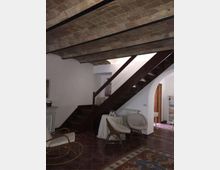 Interno di una casa con soffitto a travi e voltine in mattoni e pavimento in cotto, dominato da una scala in legno scuro che sale lungo la parete. Sotto la scala c’è un piccolo angolo con poltrone in vimini e tavolino rotondo, accanto a un arco che conduce a un’altra stanza.