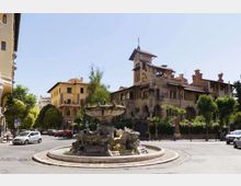 Rotatoria urbana con grande fontana in pietra al centro, circondata da strade con auto parcheggiate e alberi. Sullo sfondo si vede una villa storica in stile liberty con torre, archi e tetti in tegole, affiancata da edifici residenziali.