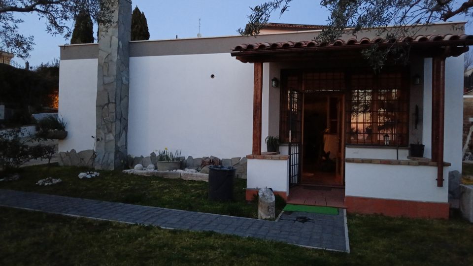 Facciata di una casa a un piano con pareti bianche e tetto in coppi, con portico d’ingresso sorretto da colonne in legno e grandi finestre con inferriate. Davanti c’è un piccolo giardino con prato, aiuole di pietre e un vialetto pavimentato che conduce alla porta, con un alto camino rivestito in pietra sul lato sinistro.
