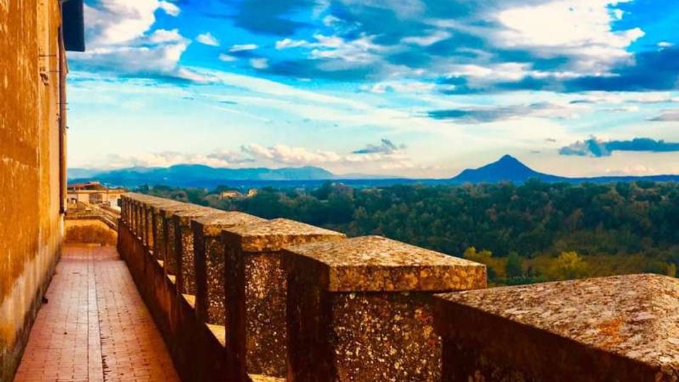 Terrazza panoramica di un edificio in pietra con lungo camminamento in mattoni e parapetto massiccio, affacciata su una vasta area boscosa. Sullo sfondo si vedono colline e una montagna isolata sotto un cielo ampio con nuvole.