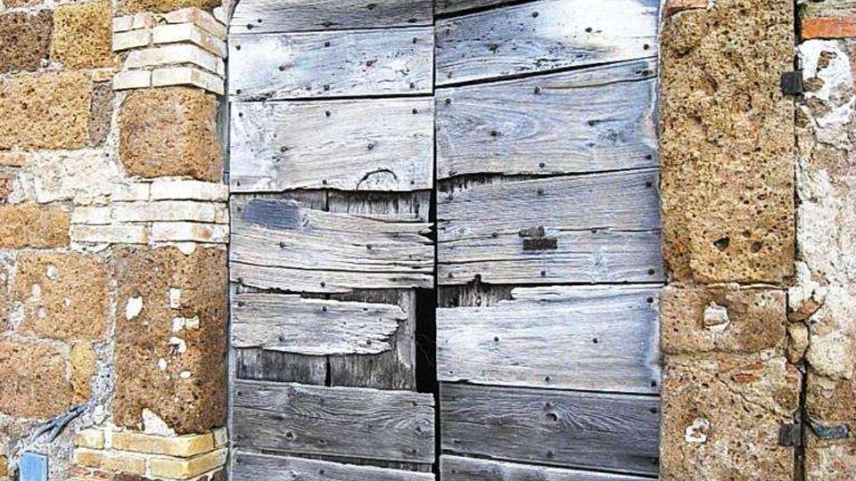 Porta ad arco in legno molto usurato e parzialmente rotto, inserita in una muratura in pietra con cornice in blocchi e piccoli inserti in mattoni. Sopra la porta c’è una grata in ferro battuto decorativa, con pavimentazione in pietra e ciottoli davanti all’ingresso.