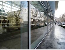 Marciapiede lungo un edificio moderno con grande facciata vetrata riflettente e pensilina in vetro e metallo. Accanto corre una strada con auto e alberi spogli, con edifici sullo sfondo.