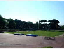 Pista di atletica all’aperto con ampio prato centrale e pedane per salto in alto con materassi blu. Sullo sfondo si vedono alberi alti e una linea di verde che delimita l’area sportiva.