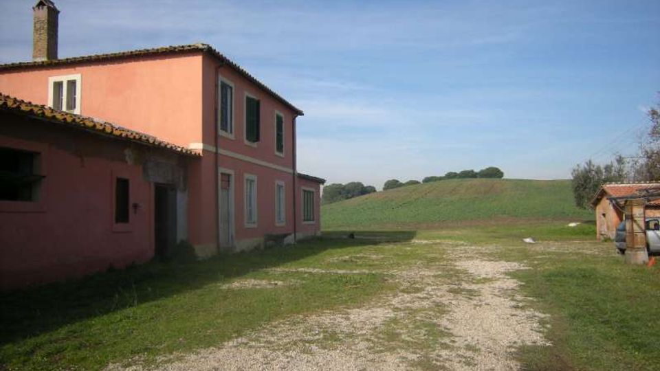 Cascina di campagna con edificio intonacato rosa a due piani, tetto in coppi e persiane scure, affacciata su un cortile in ghiaia e prato. Sullo sfondo si vedono campi coltivati ondulati e un piccolo annesso rustico ai margini della proprietà.