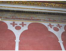Dettaglio di una parete interna decorata con archi stilizzati rossi e colonne bianche, sormontata da fasce ornamentali policrome a motivi geometrici e iscrizioni. La parte superiore mostra cornici e fregi con mosaico o pittura decorativa.