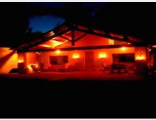 Casa a un piano vista di notte con ampio portico coperto da tetto a falde e travi a vista, illuminata da luci calde sulle pareti. Davanti si vede un patio con sedie e tavolo, circondato dal buio del giardino esterno.